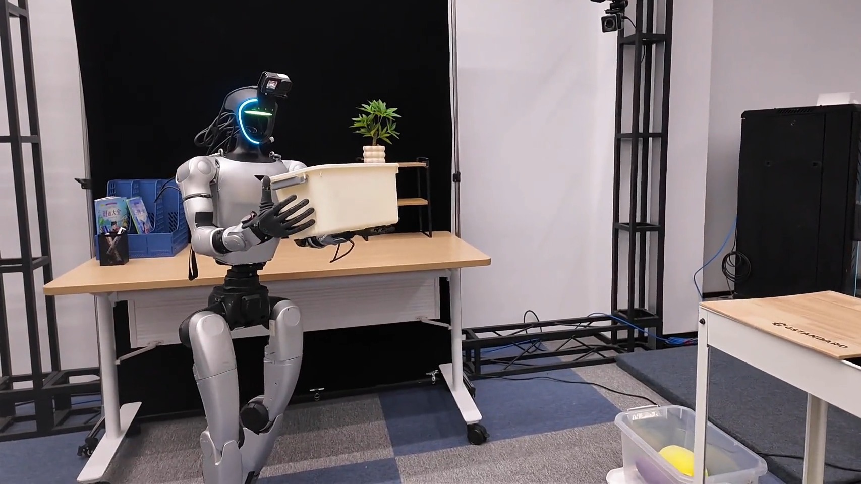 Silver humanoid robot with a glowing visor holding a white plastic box in both hands beside a wooden desk