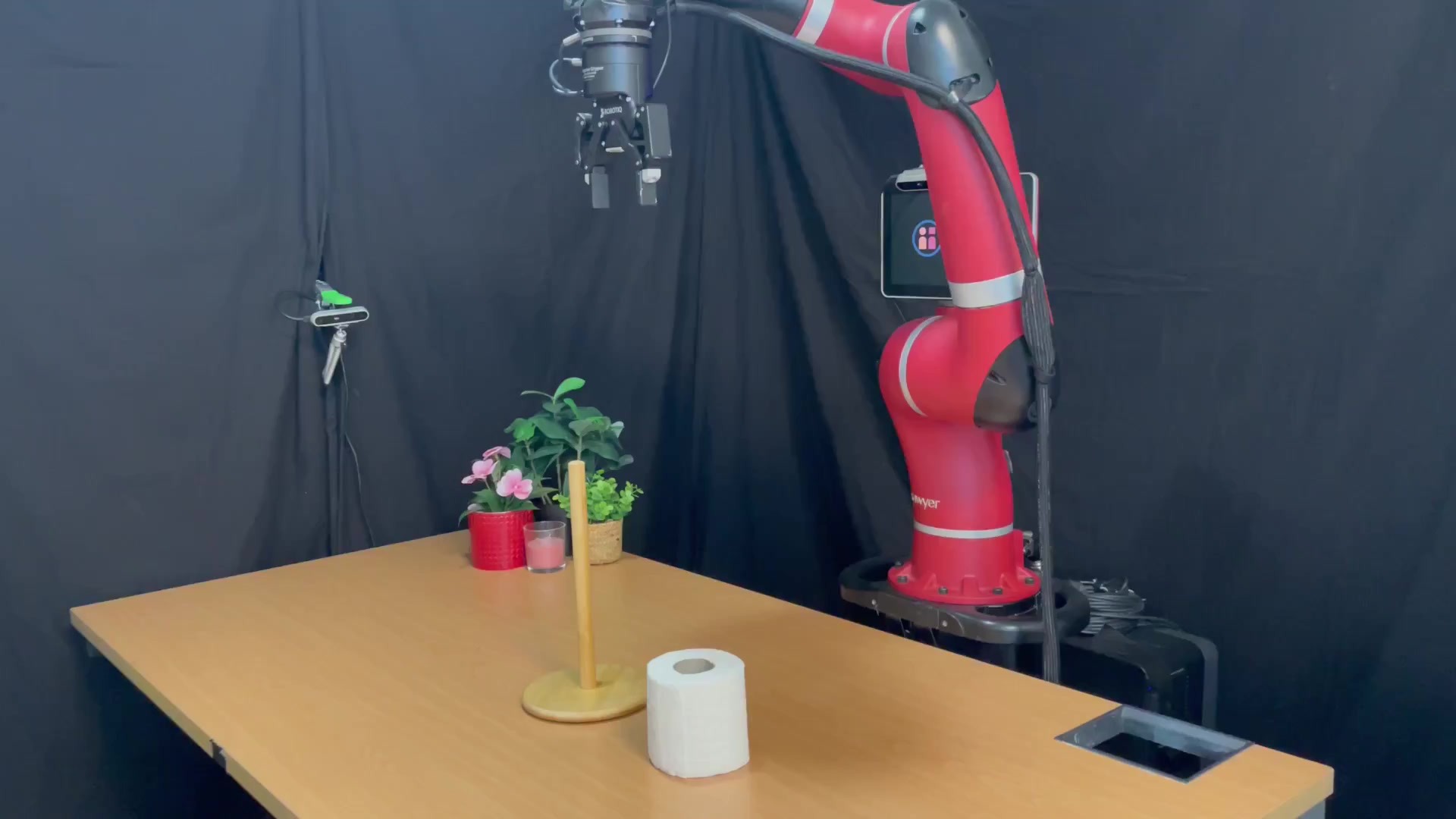 Red one-armed robot with a black gripper above a wooden desk with a roll of toilet paper, a wooden peg and potted flowers